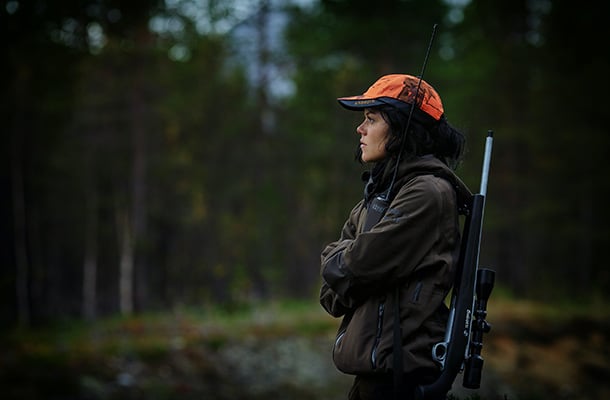 En kvinnelig jeger i skogen kledd i jaktklær og oransje caps, bærer rifle på ryggen.