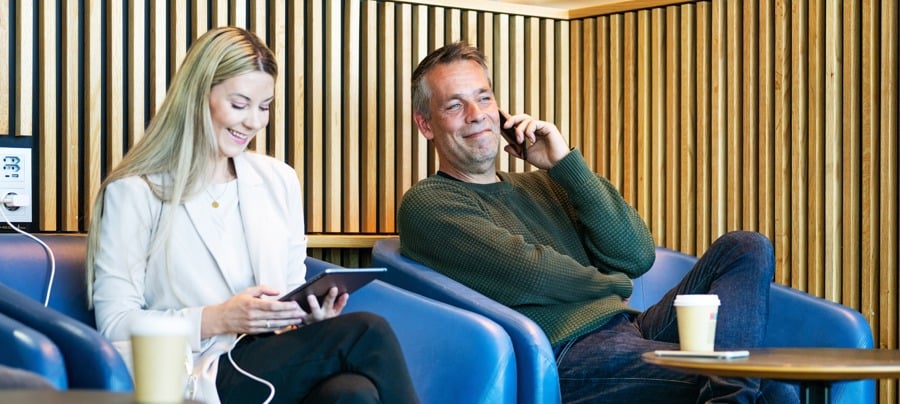 To personer slapper av i en moderne kafé. En kvinne ser på et nettbrett og en mann snakker i telefon. Begge sitter i blå stoler med kaffekopper på bordet foran seg.