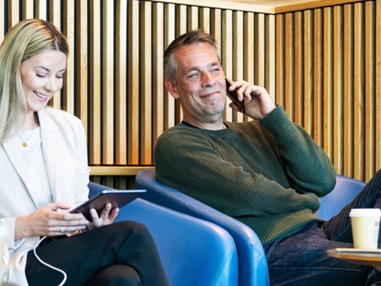 To personar slappar av i ein moderne kafé. Ei kvinne ser på eit nettbrett og ein mann snakkar i telefon. Begge sit i blå stolar med kaffikoppar på bordet framfor seg.