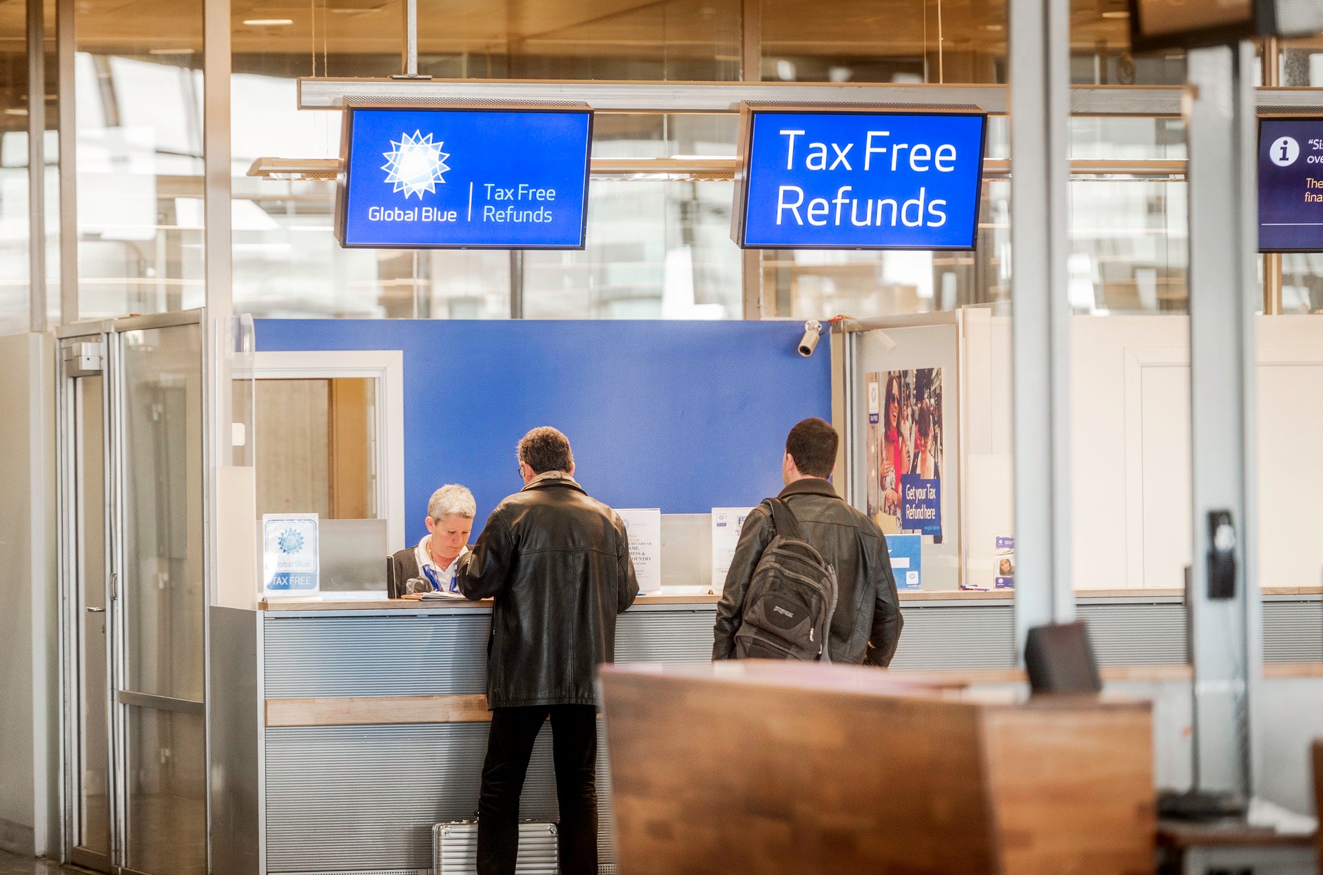Personer ved en Global Blue tax-free-refusjonsdisk på en flyplass. To menn står ved skranken under et blått skilt som viser "Tax Free Refunds".