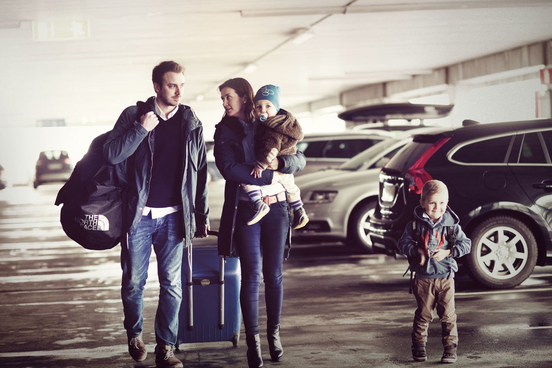 Family of four with luggage in a parking garage, ready for travel. Parents and children stand among cars with suitcases and backpacks.