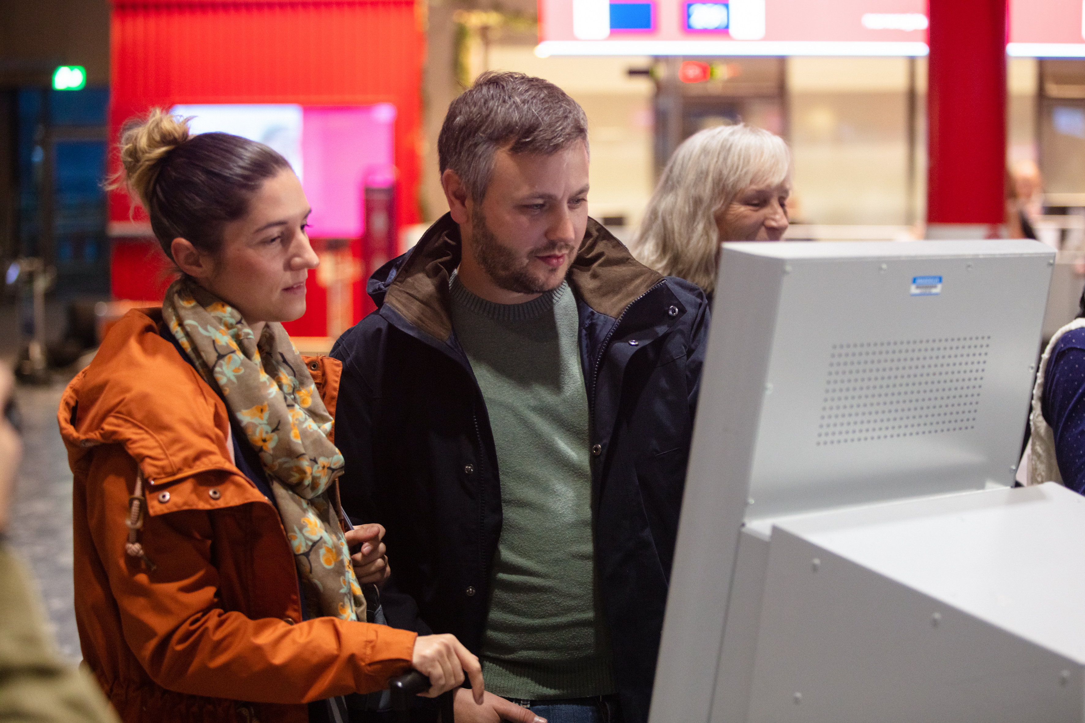 To personer sjekker inn på en digital skjerm på en flyplass. De er kledd i høstjakker.