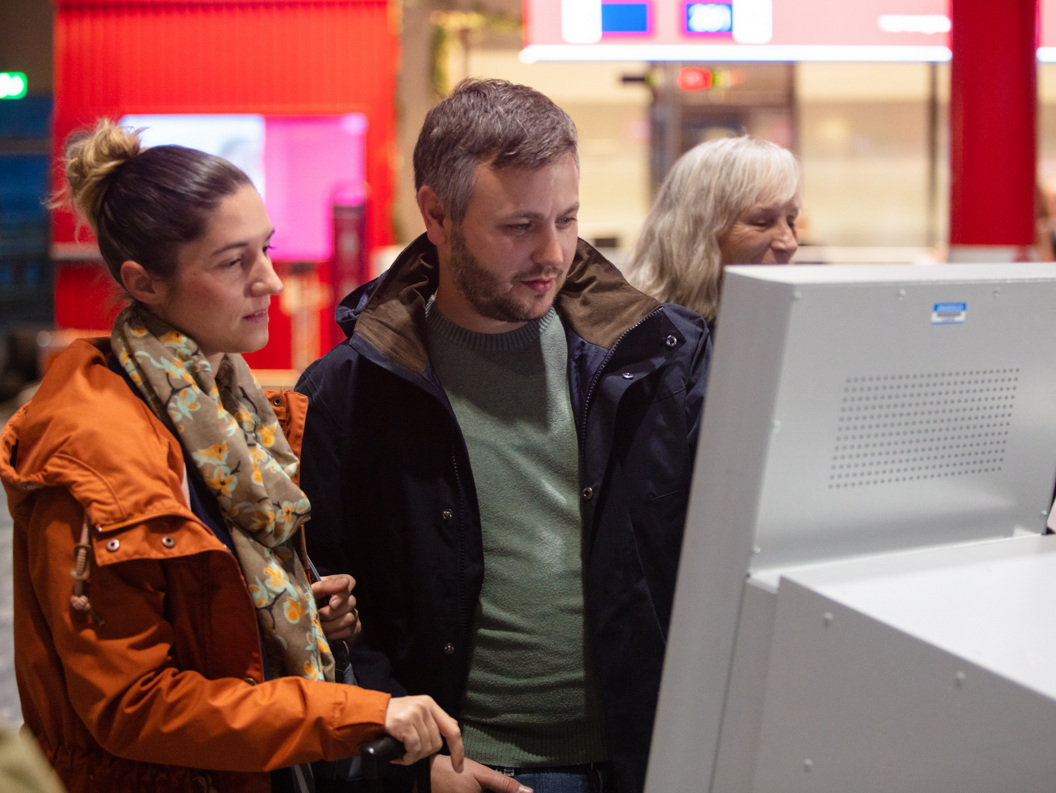 To personar sjekkar inn på ein digital skjerm på ein flyplass. Dei er kledde i haustjakker.