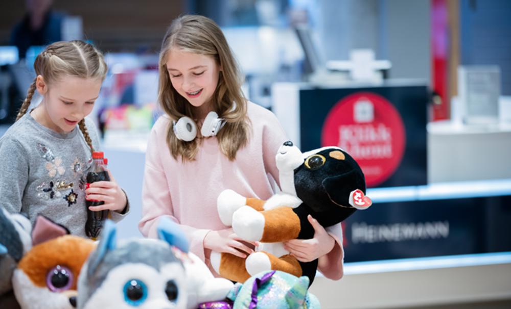 Two children are looking at stuffed animals in a store. One of them is holding a stuffed dog, while the other has a soda bottle.