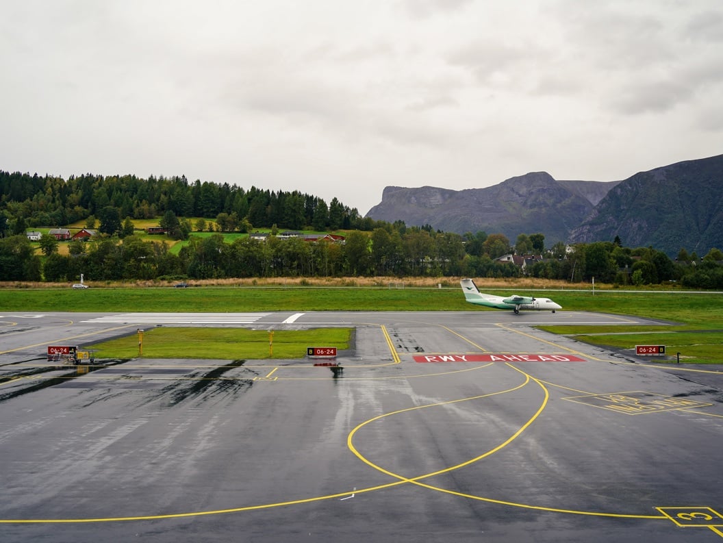 Småfly på rullebanen ved ein flyplass omgitt av frodig natur, med skogkledde åsar og fjell i bakgrunnen.