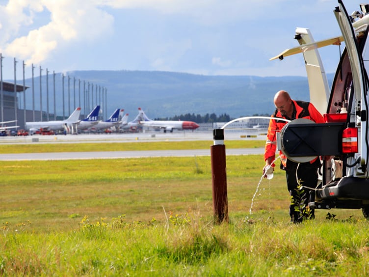 Mann ved open varebil utfører kontroll på flyplass med fly og terminal i bakgrunnen