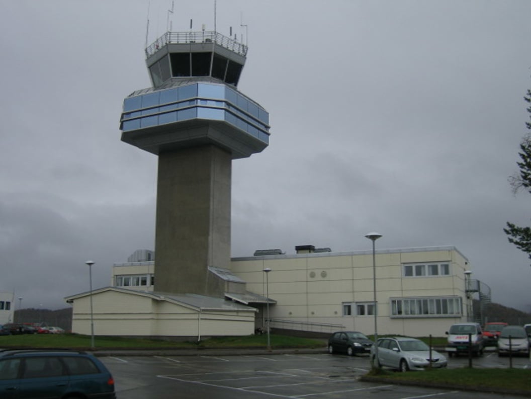Control tower at Bardufoss Airport surrounded by cloudy skies and parked cars.