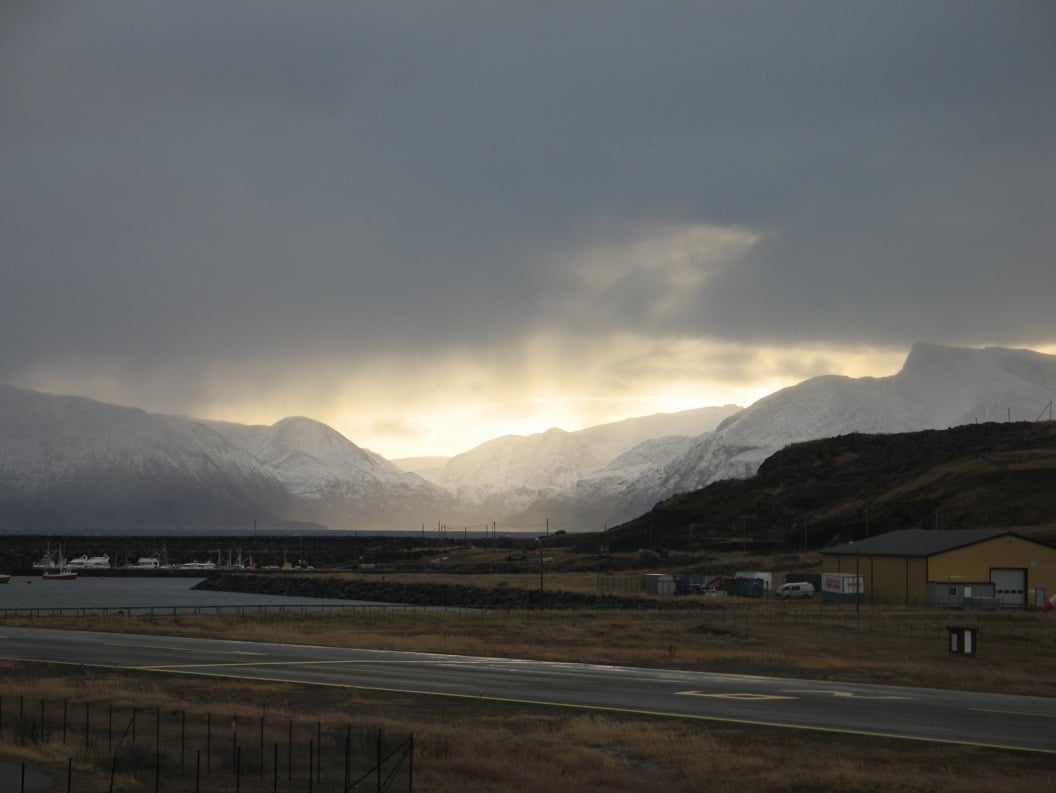 Landskap av snødekte fjell under dramatiske skyer og lysstråler ved Hasvik lufthavn, Norge.