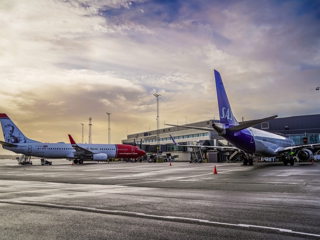 To fly på ein flyplass i solnedgangen, eit Norwegian-fly og eit SAS-fly parkert ved ein terminal, med eit moderne terminalbygg i bakgrunnen.