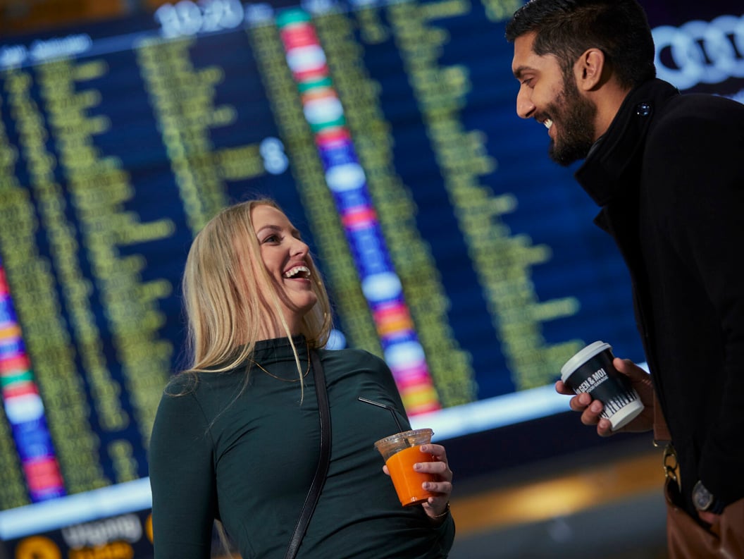 To personer ler og holder drinker foran en stor avgangstavle på flyplassen som viser flyinformasjon