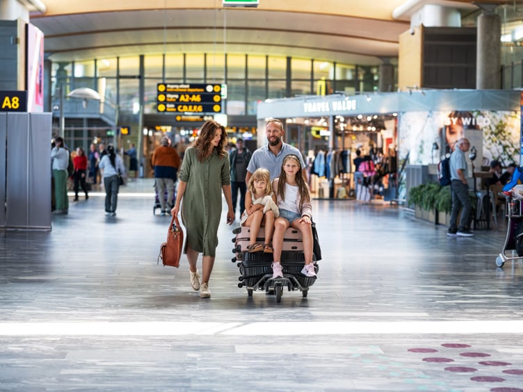 Familie med bagasjevogn på flyplass, blant reisebutikker og avganger.