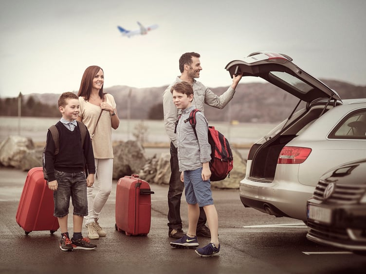 Familie på fire ved bil parkeringsplass, med kofferter og rygsekker klare for reise.