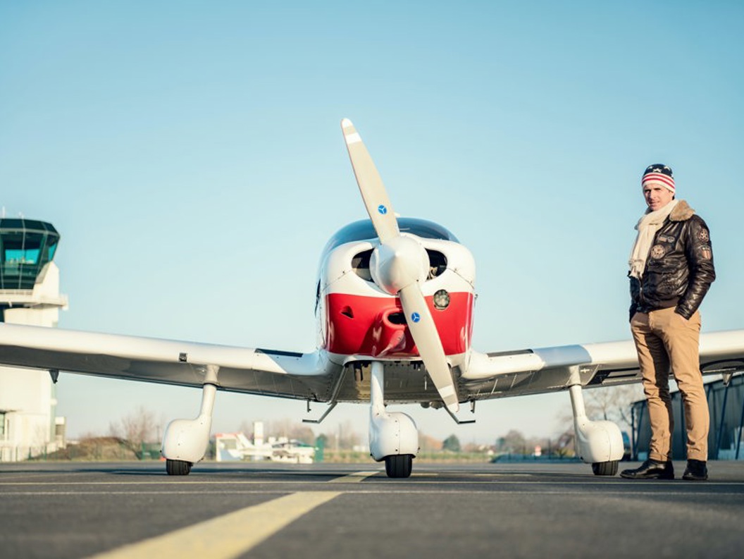 Light propeller plane at the airport with a control tower in the background, a man dressed in winter clothing standing next to the plane.