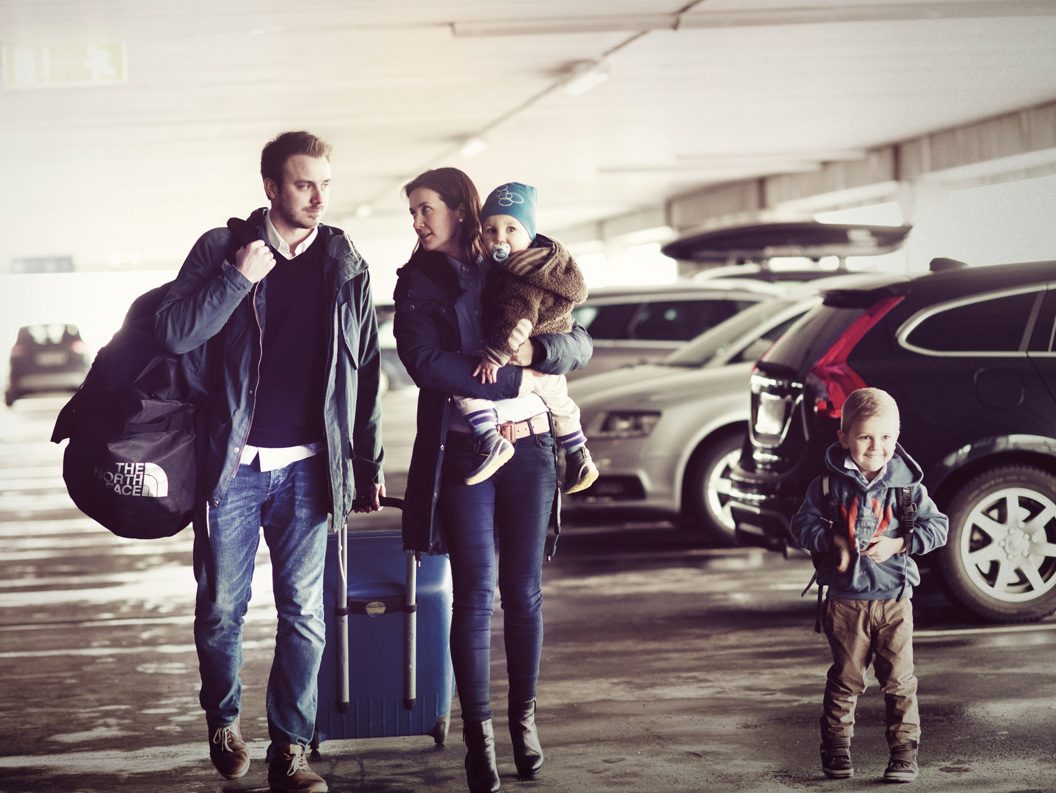Familie på fire med bagasje i parkeringshus, klare for reise. Foreldre og barn står blant biler med kofferter og ryggsekker.