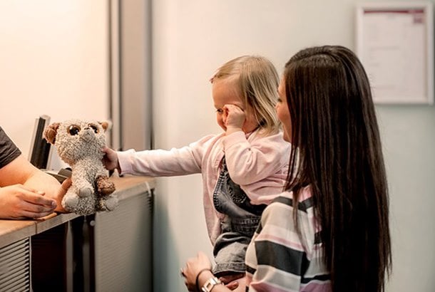 A lady with long brown hair stands with a little girl on her arm in front of a counter where the girl gets back a teddy bear she has forgotten on the plane or similar.
