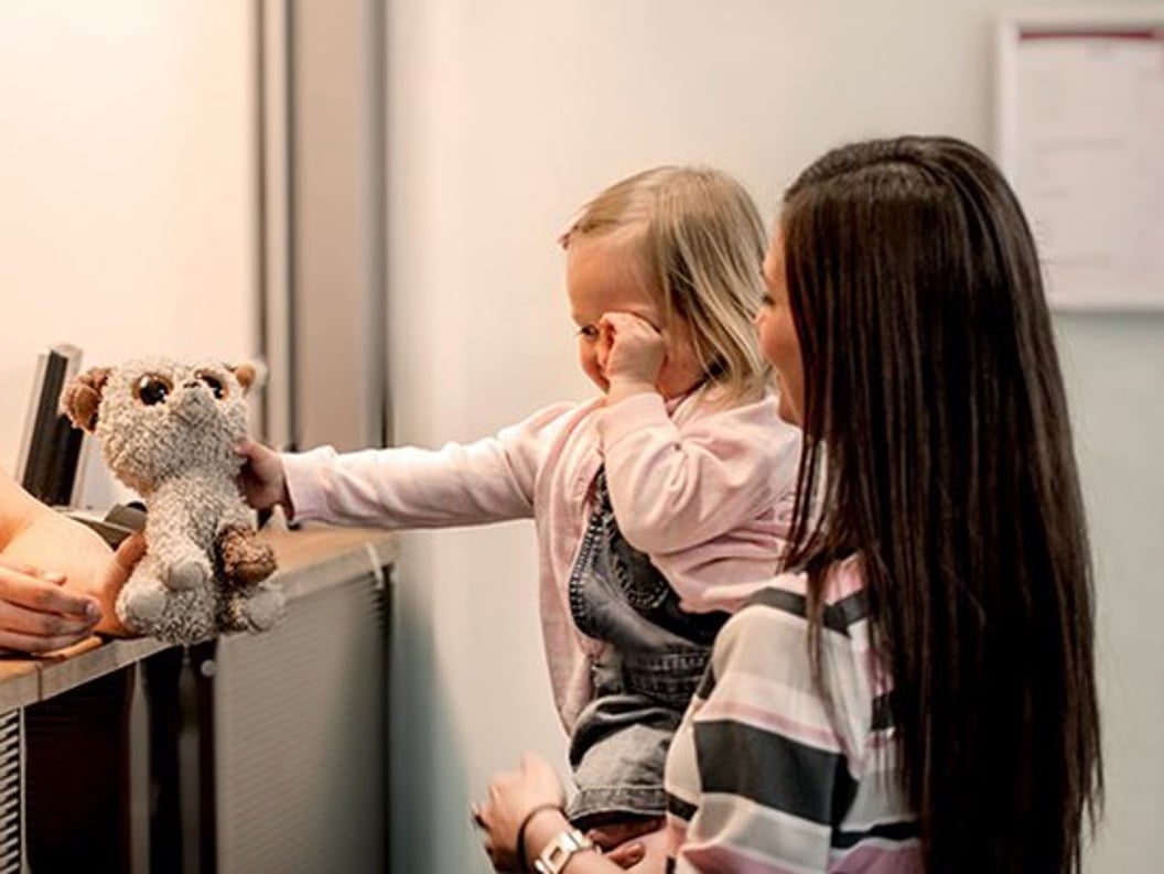 A lady with long brown hair stands with a little girl on her arm in front of a counter where the girl gets back a teddy bear she has forgotten on the plane or similar.