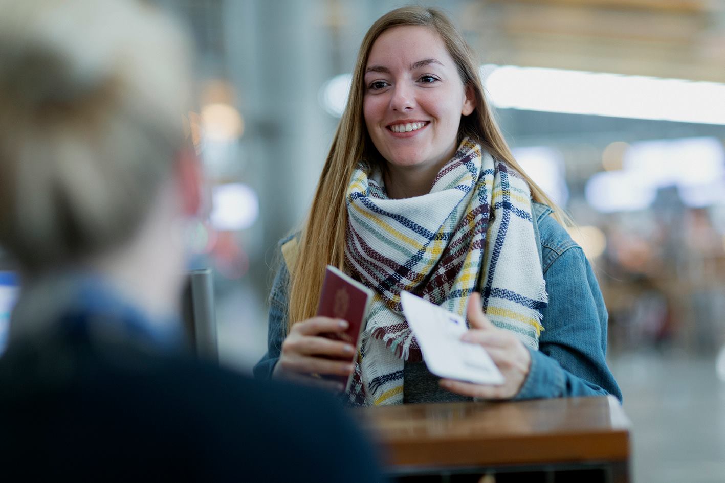 A lady with long, semi-dark hair is standing in front of a counter at the airport terminal with her passport in her hand.