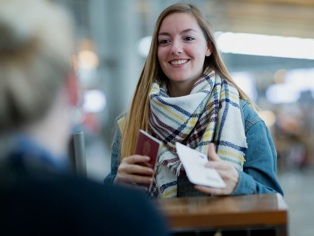 En dame med langt halvmørkt står foran en skranke på flyplassterminalen med passet sitt i hånden.
