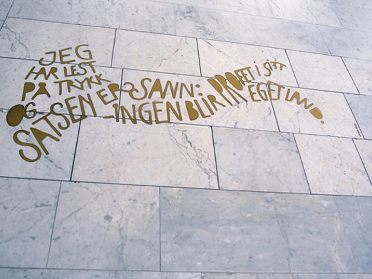 Tekst på gulv laget i gullbokstaver på marmorfliser, med teksten: "JEG HAR LEST PÅ TRYKK OG SAVN ER SATSEN: INGEN BLIR DRØFTET SÅ PROFT I SITT EGET LAND