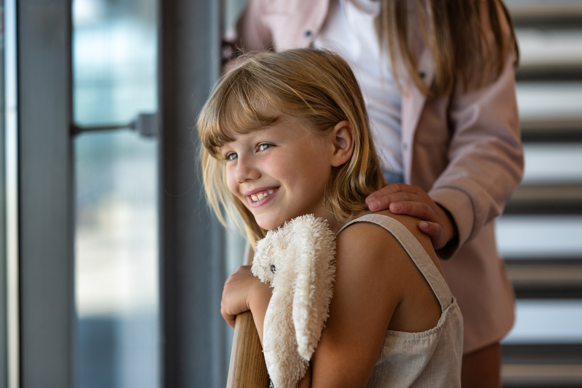 Girl with blonde hair smiles while holding a soft teddy bear. An adult's hand gently rests on her shoulder. Natural light from a window in the background.