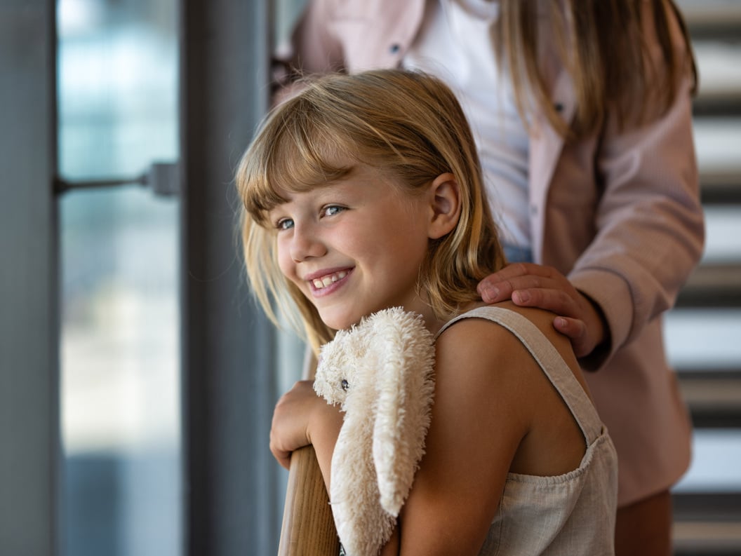 Girl with blonde hair smiles while holding a soft teddy bear. An adult's hand gently rests on her shoulder. Natural light from a window in the background.