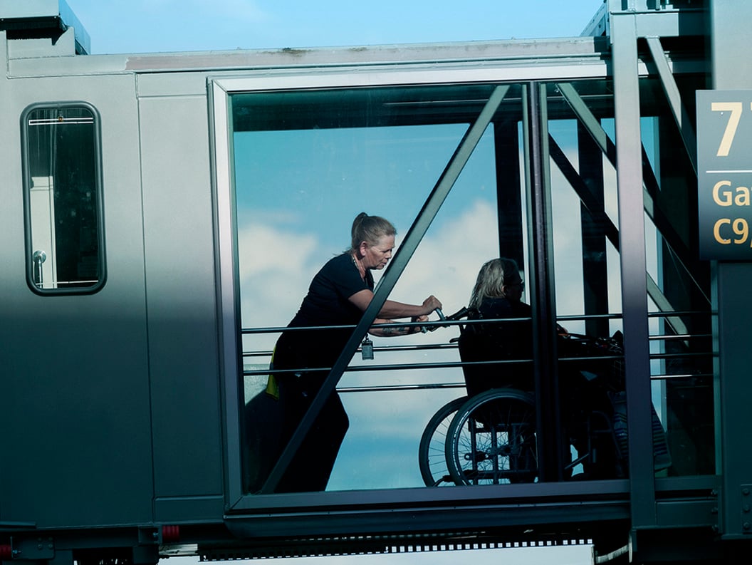 Dame som assisterer en annen dame i rullestol ut av flyet. Vi ser de gjennom vindusruten i overgangen mellom flyet og terminalen.