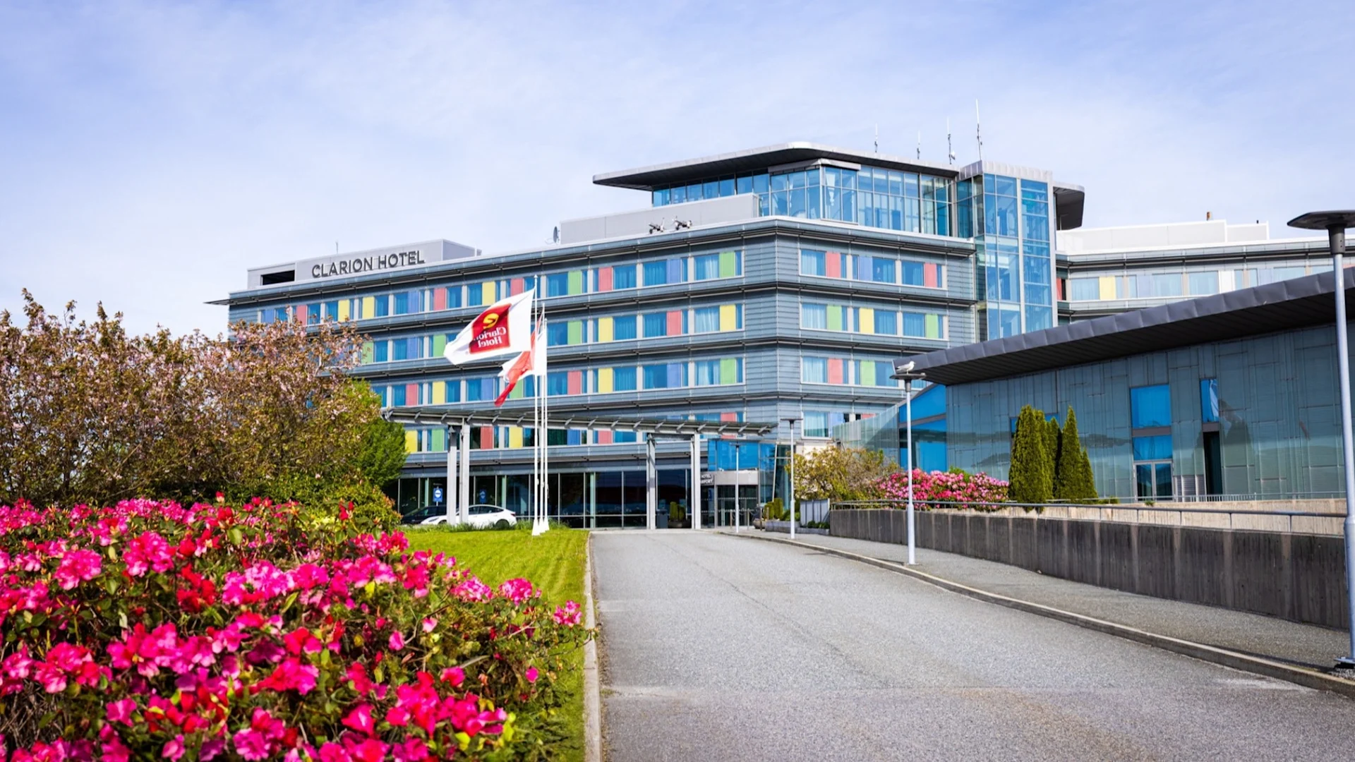 Moderne fasade av Clarion Hotel omgitt av frodige blomar og tre under ein klar himmel.