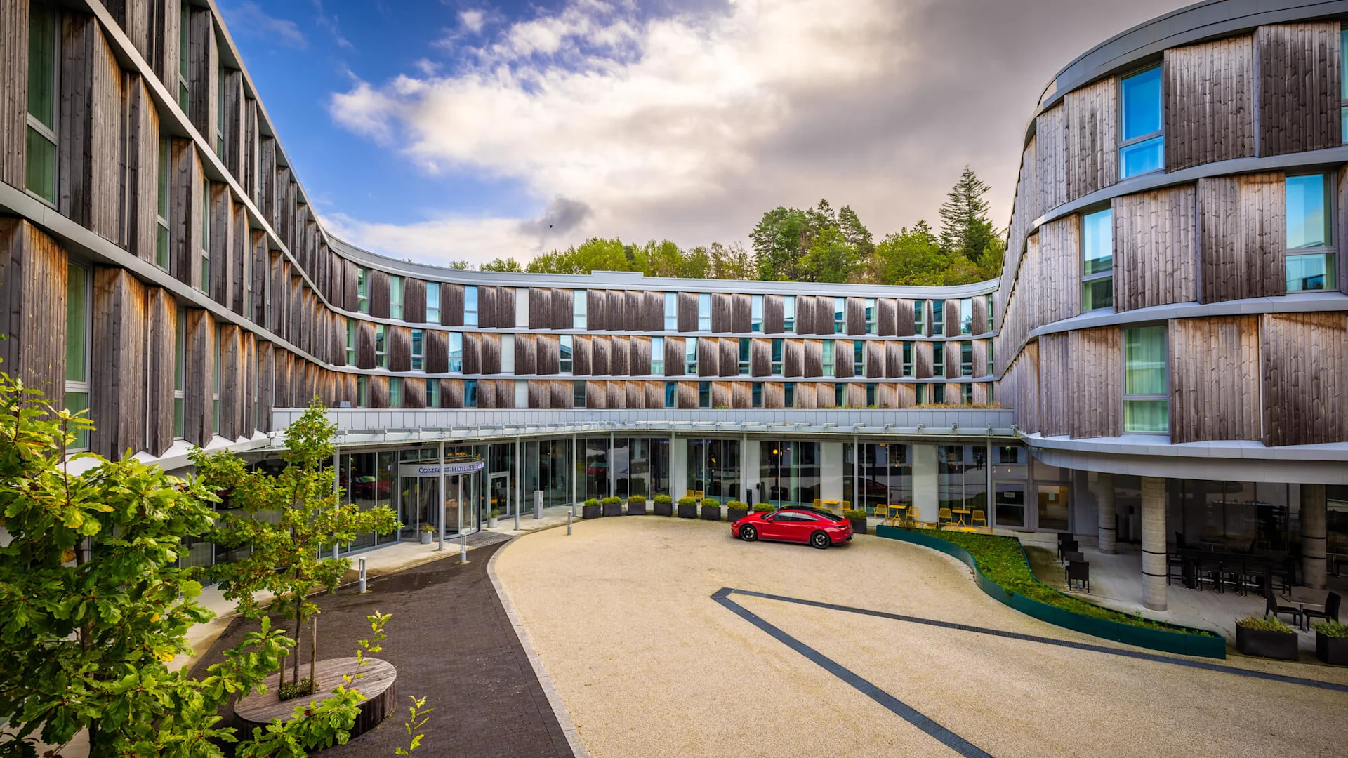 Moderne hotellfasade med bogeforma design, store vindauge, og ein raud bil parkert framfor hovudinngangen, omgitt av grøntområde og delvis overskya himmel.