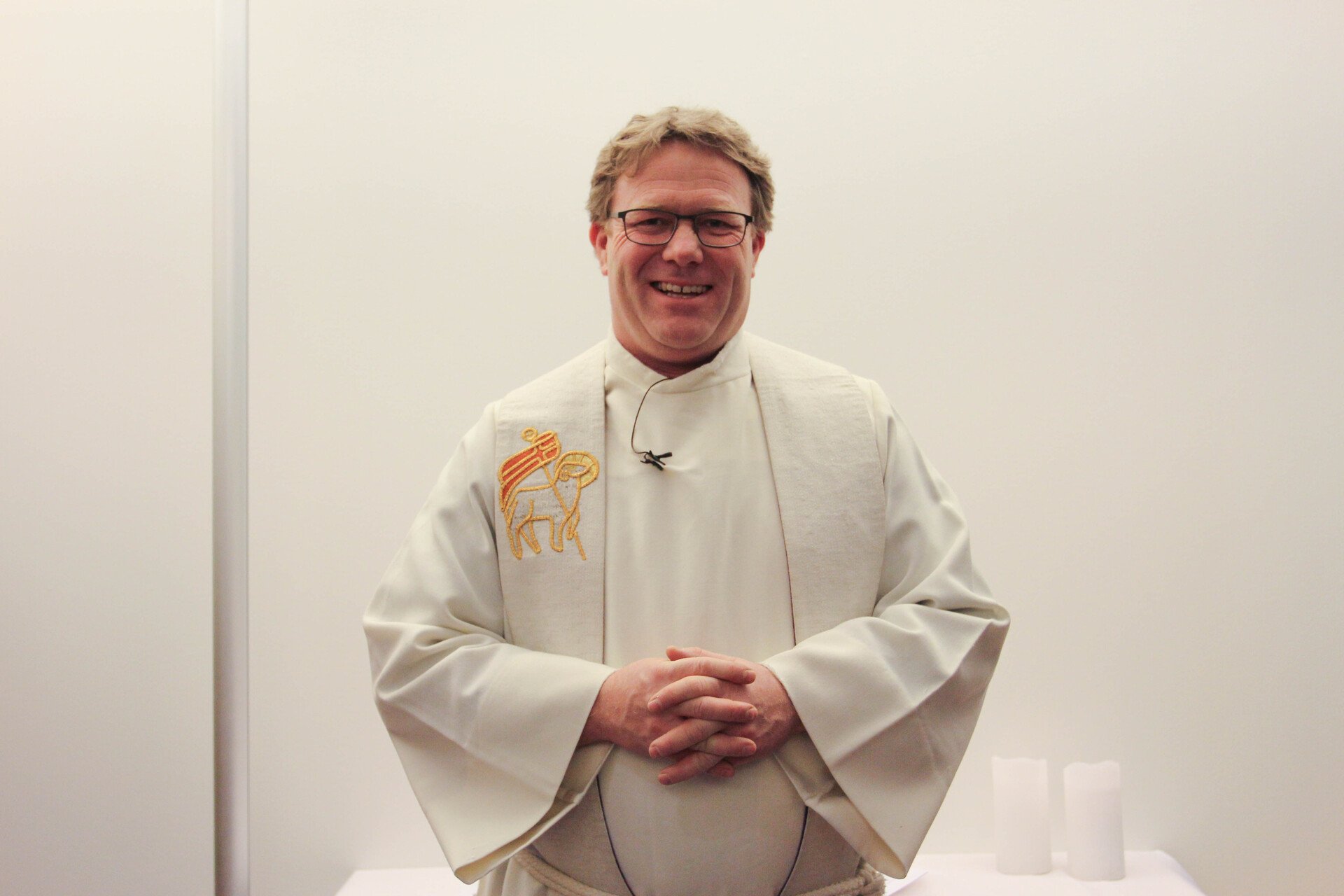 En smilende prest i hvit kappe står foran en lys vegg, med et brodert symbol på venstre side av kappen.