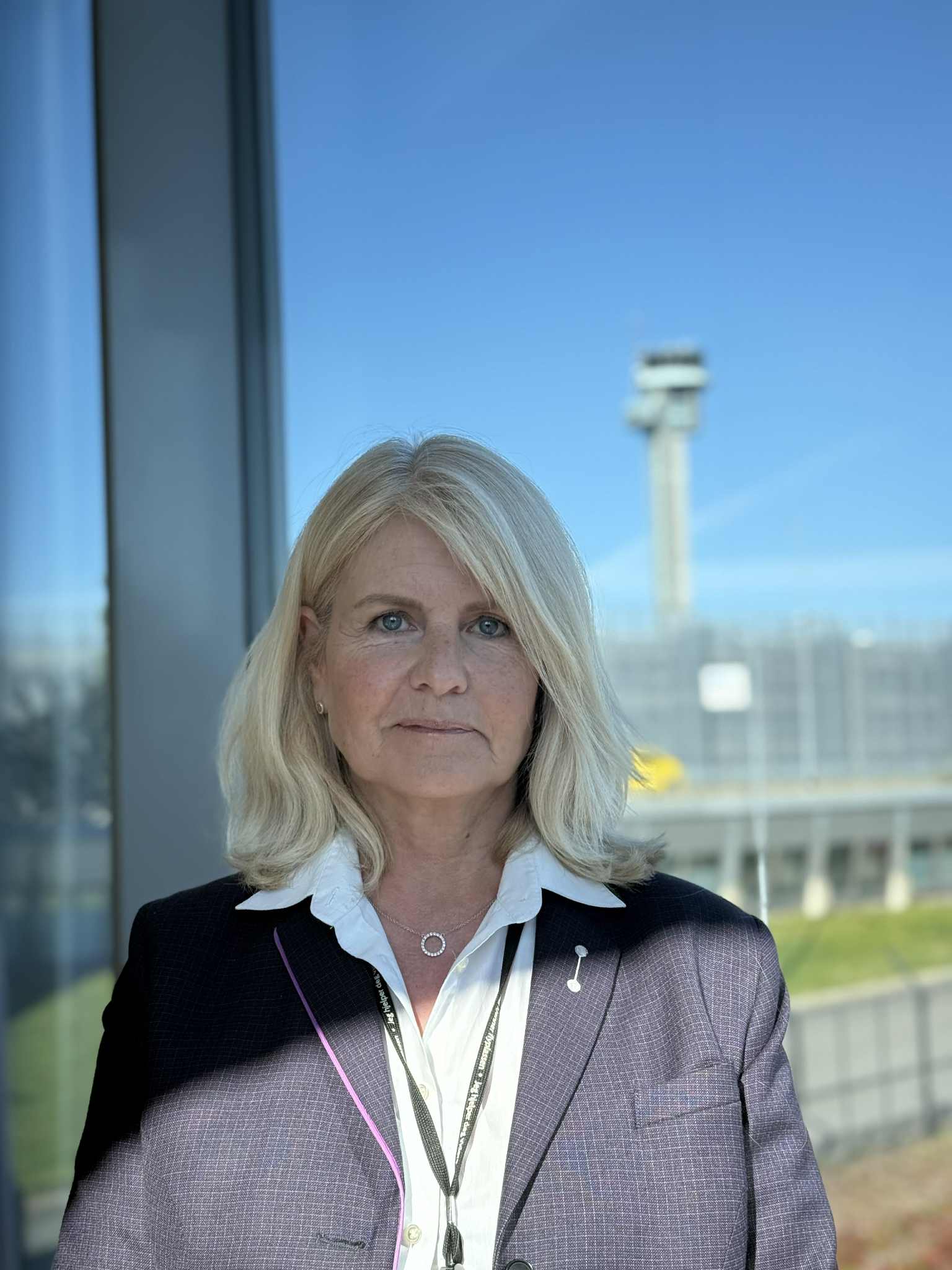 Kvinne i dressjakke med hvit skjorte står foran et glassvindu med utsikt over en flyplassterminal og kontrolltårn i bakgrunnen.