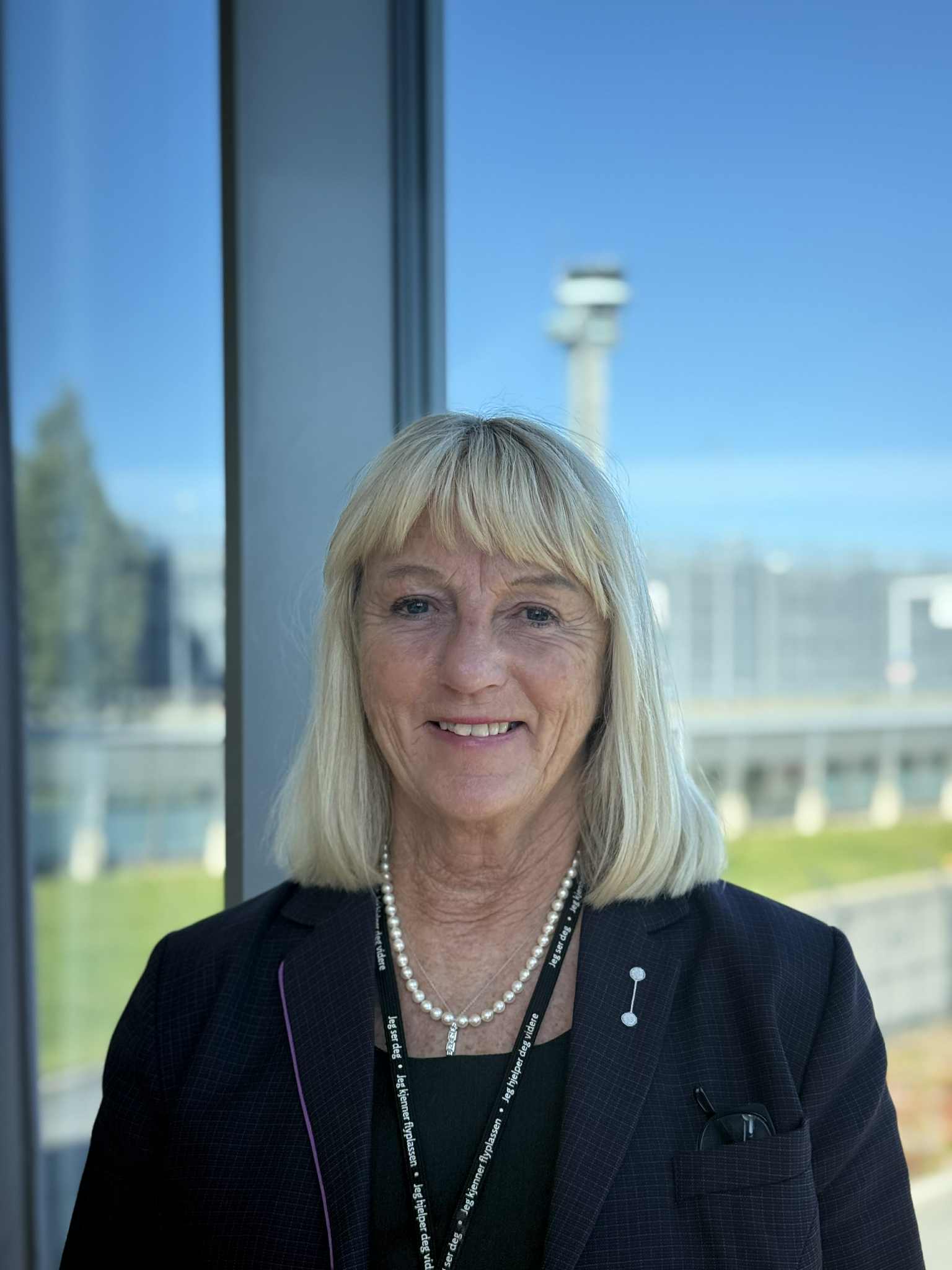 Kvinne med blondt hår i en mørk blazer smiler foran et vindu med utsikt over et åpent landskap og en uklar struktur i bakgrunnen.
