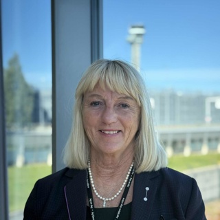 Woman with blonde hair in a dark blazer smiling in front of a window overlooking an open landscape and a blurry structure in the background.