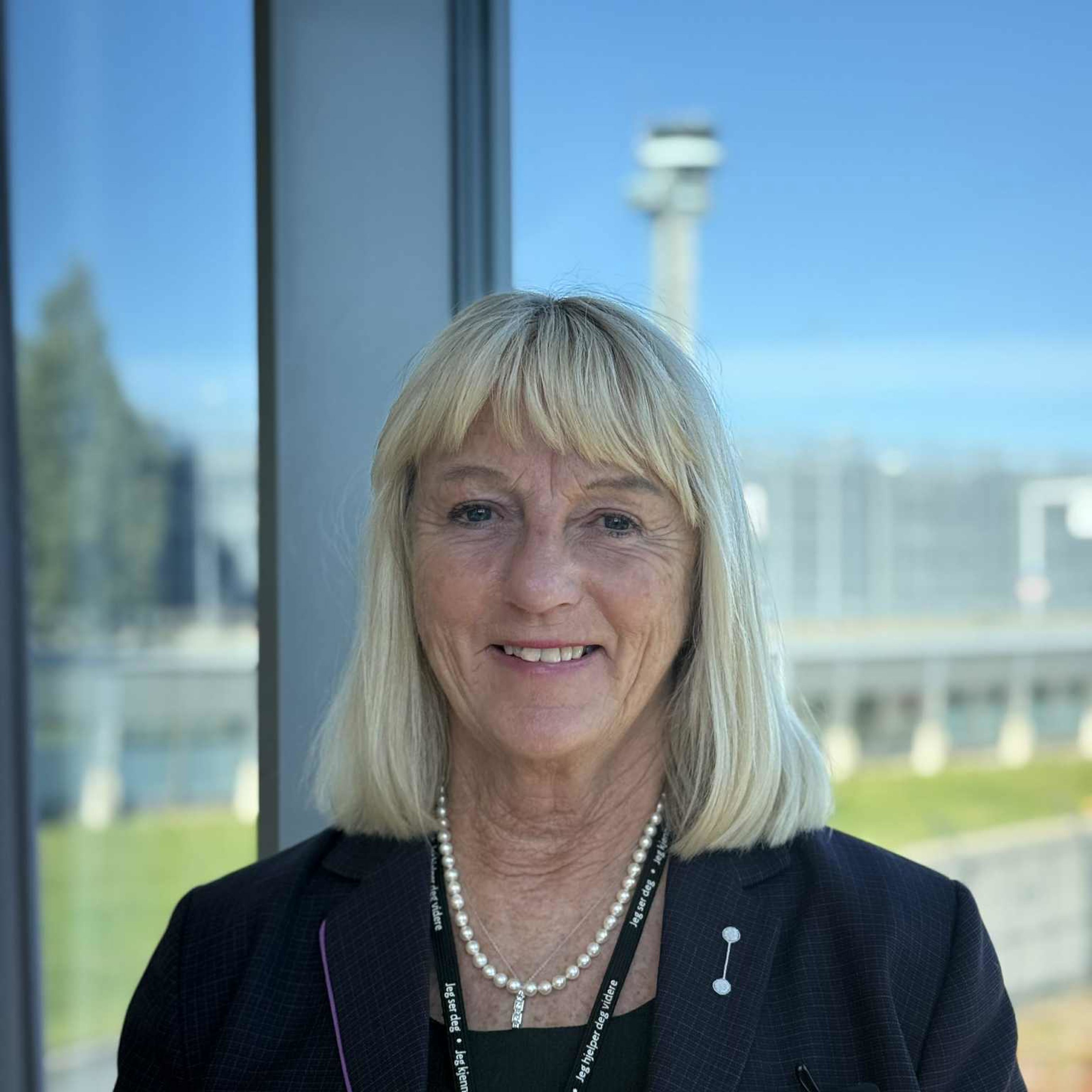 Kvinne med blondt hår i en mørk blazer smiler foran et vindu med utsikt over et åpent landskap og en uklar struktur i bakgrunnen.