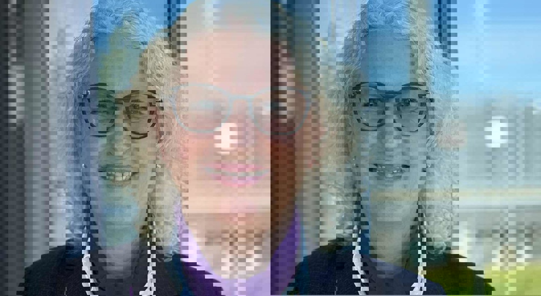 Woman with light curly hair and glasses stands by a window wearing a dark jacket and shirt, in front of an airport background with a clear blue sky.