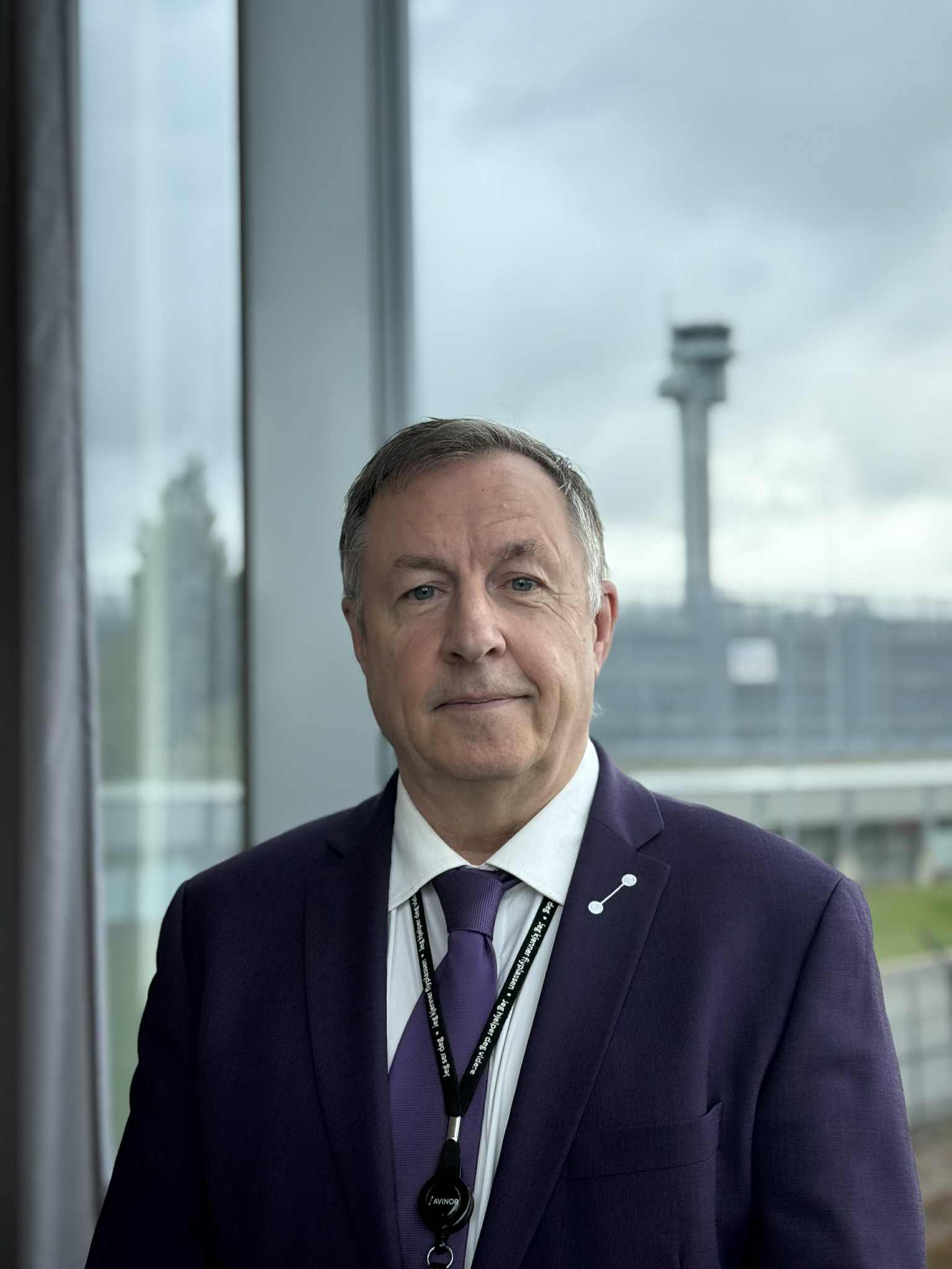 En mann i dress står foran et stort vindu med utsikt mot en flyplassterminal og kontrolltårn i bakgrunnen.
