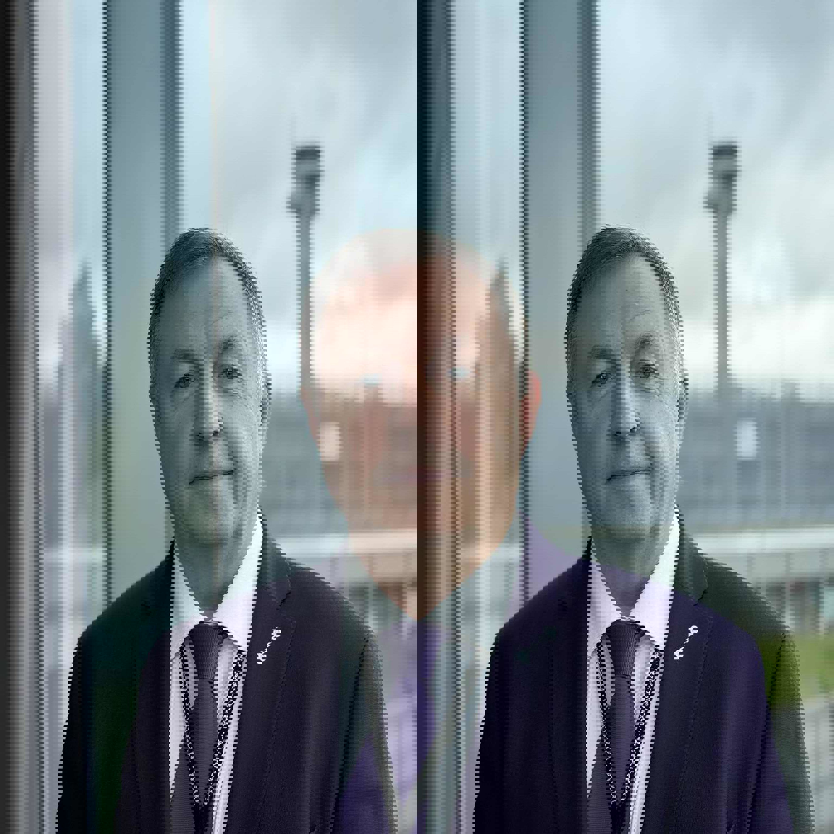 En mann i dress står foran et stort vindu med utsikt mot en flyplassterminal og kontrolltårn i bakgrunnen.