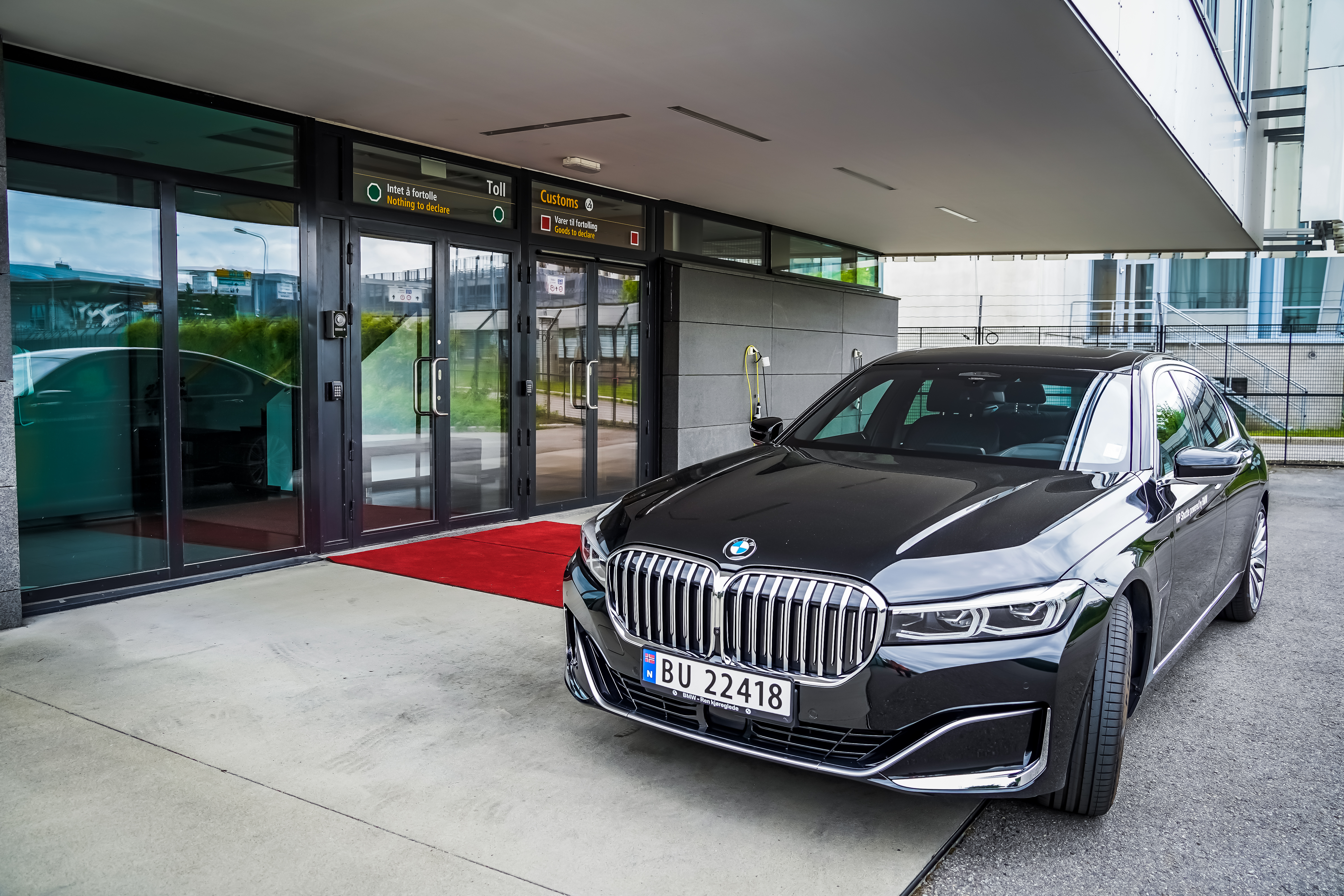 Svart BMW parkert foran en tollinnsjekking ved en bygning med glassdører, rød løper langs inngangen.