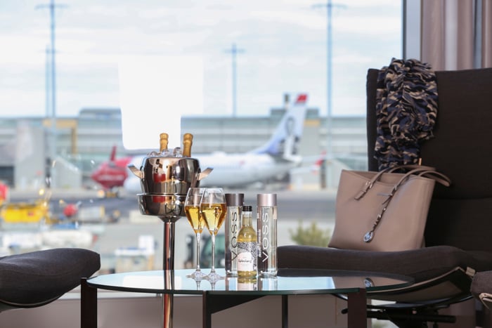 Luksuriøs flyplasslounge med utsikt over rullebane, sjampanje og Voss-flasker på bordet.