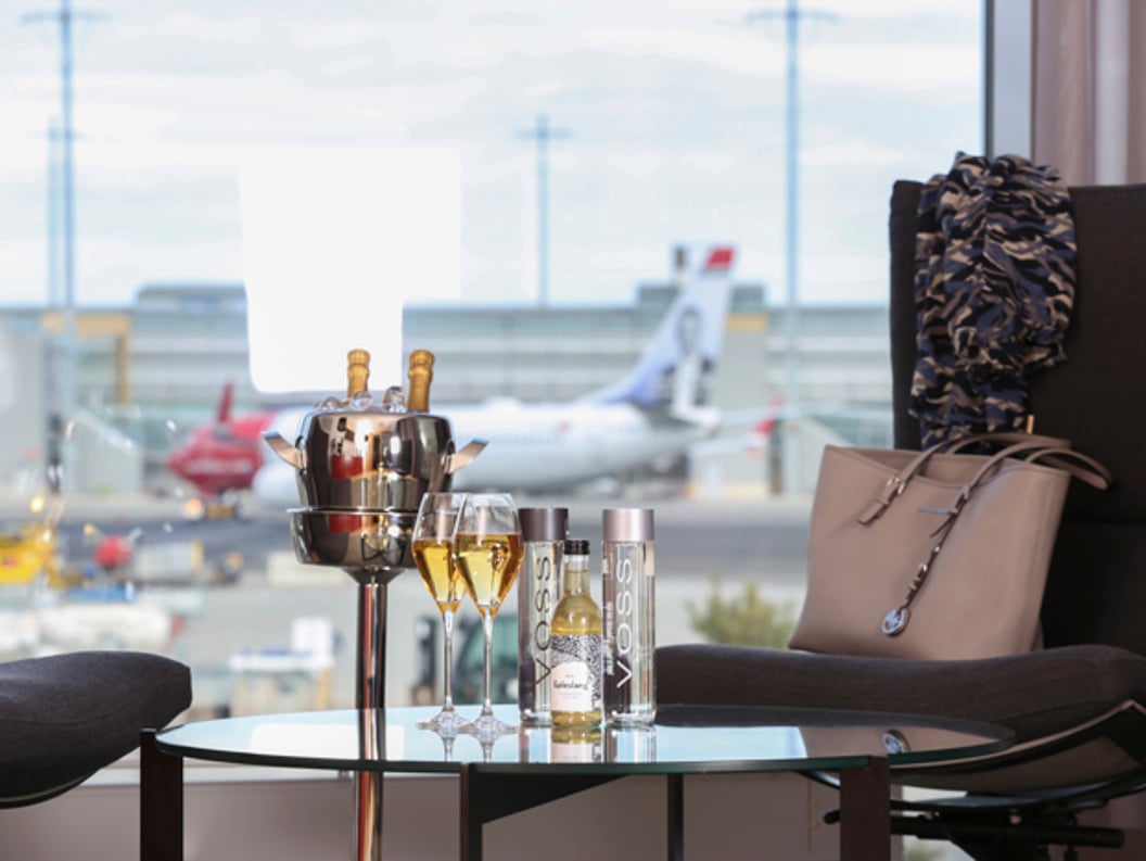 Luxury airport lounge with champagne, Voss water, and a stylish handbag on a table, overlooking an airport runway with planes in the background