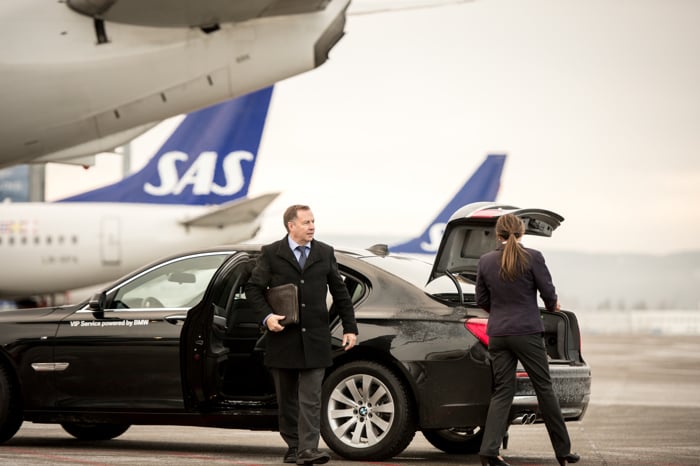Forretningsmann som stiger ut av en luksusbil ved en flyplass, med SAS-fly i bakgrunnen. En kvinne laster bagasje inn i bilens bagasjerom.