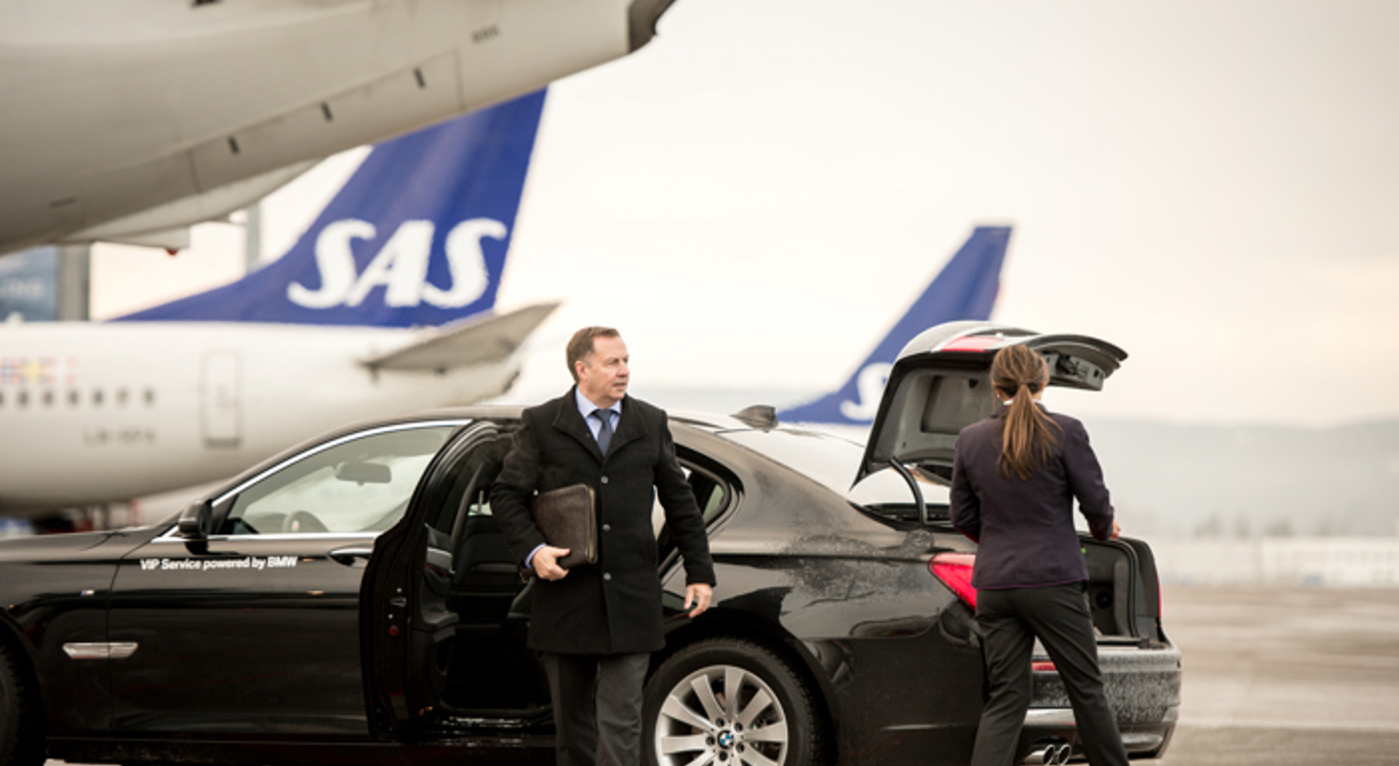 Forretningsmann som stiger ut av en luksusbil ved en flyplass, med SAS-fly i bakgrunnen. En kvinne laster bagasje inn i bilens bagasjerom.