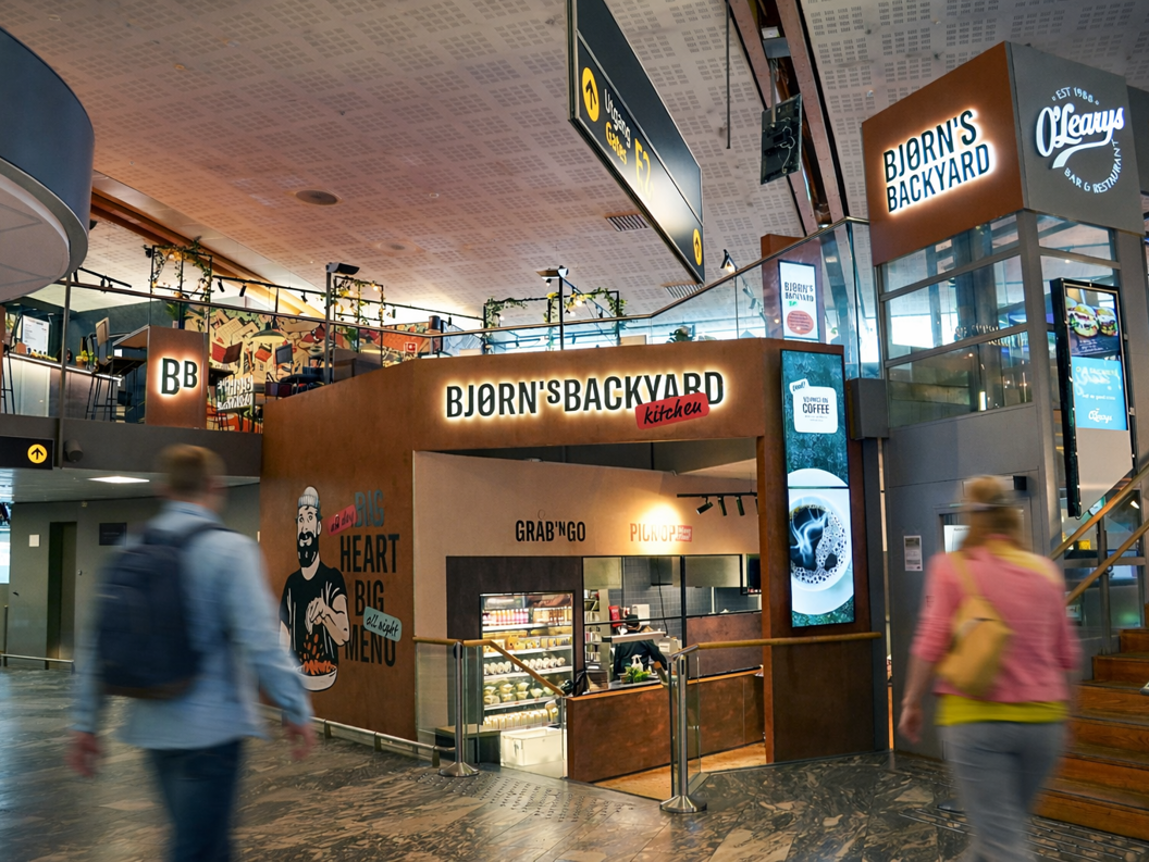Restaurant Bjørn's Backyard Kitchen i ein travel flyplass, med ein illustrasjon av ein mann som et kylling. Skiltet viser "Grab 'n Go" og "Pick Up". Tvillingane ballongar pynter inngangen