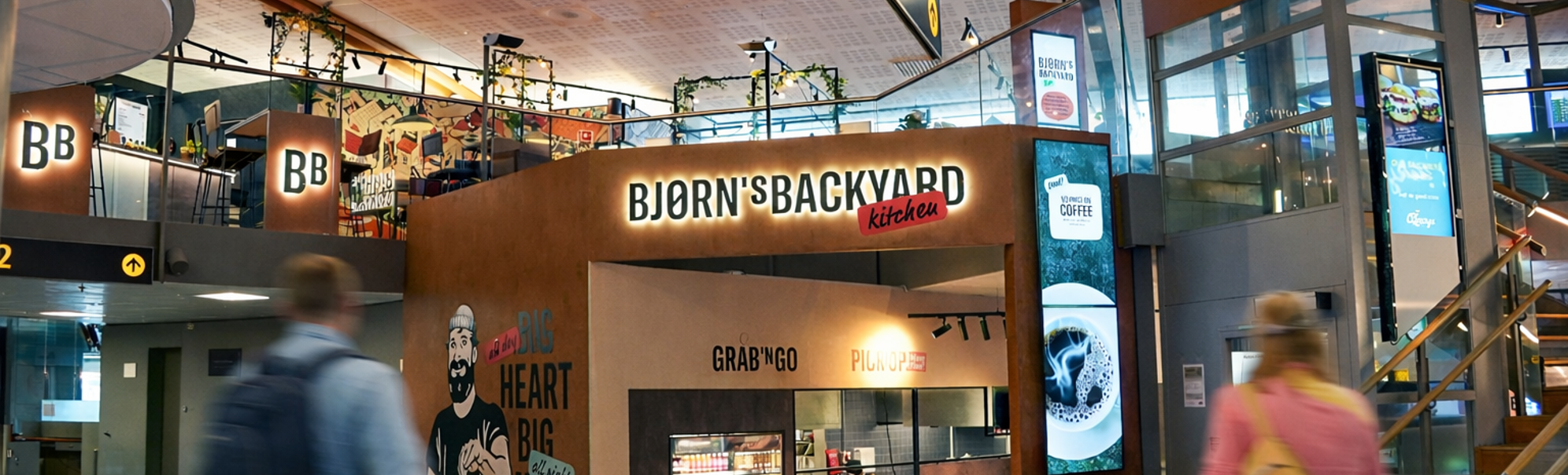 Bjorn's Backyard Kitchen restaurant entrance in a shopping mall, featuring a mural of a bearded man holding food, with slogans "Big Heart Big Menu" and "Grab 'N Go, Pick Up