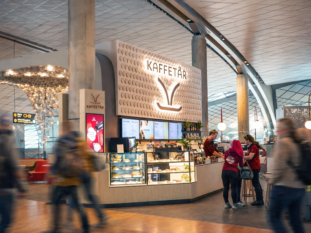 Norsk flyplasslounge med sittegruppe, dekorativ veggdesign med 'Norge', retningsskilt for porter og toaletter, og passasjerer som jobber på bærbare datamaskiner