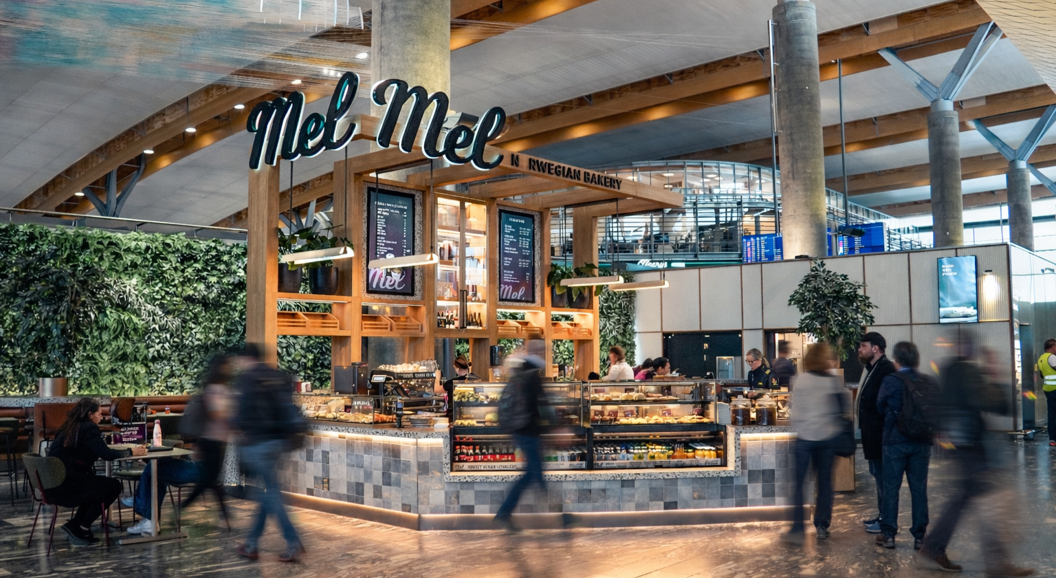 Modern bakery and café with a display of fresh pastries, customers ordering at the counter, a welcoming sign "Mel," and a barista preparing coffee. Bright, contemporary interior with a lively atmosphere