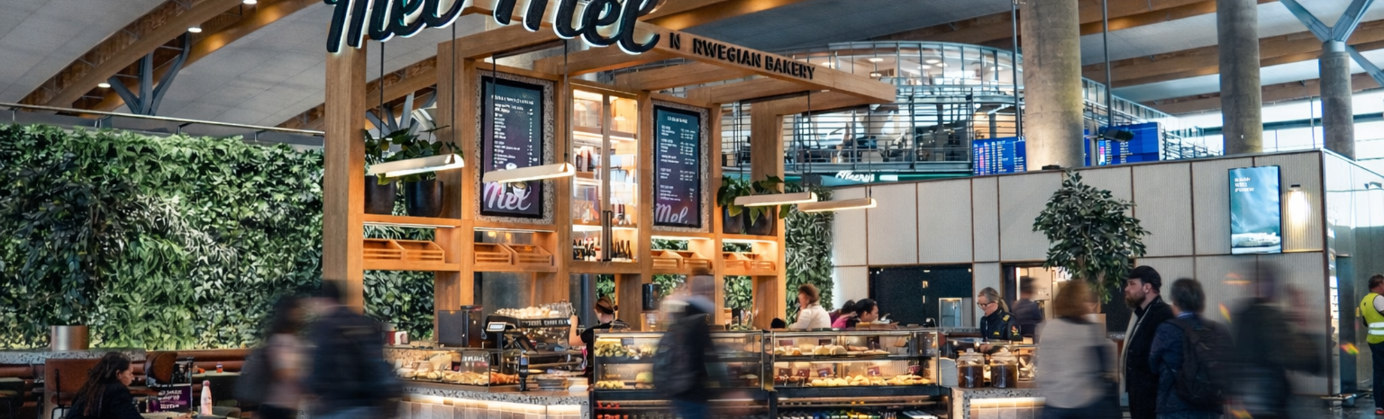 Modern bakery and café with a display of fresh pastries, customers ordering at the counter, a welcoming sign "Mel," and a barista preparing coffee. Bright, contemporary interior with a lively atmosphere