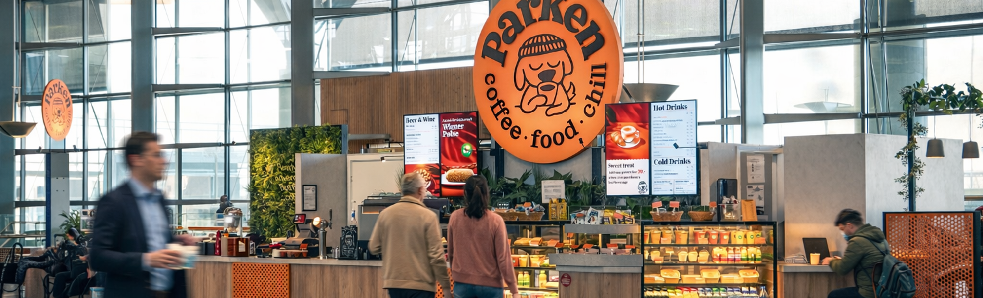 Modern airport café with large glass windows, featuring orange signage and abundant seating, offering a variety of food and beverages