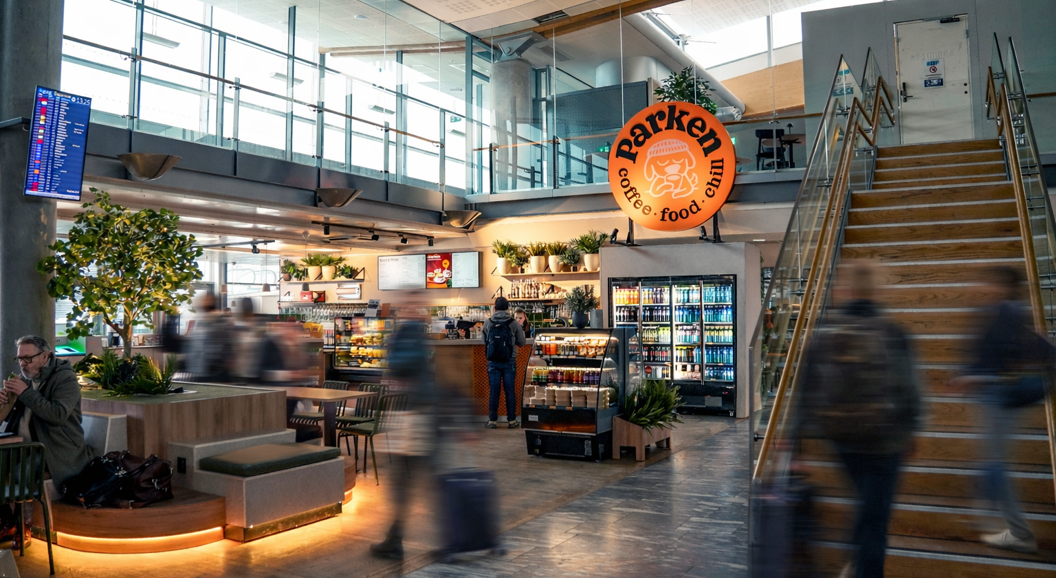 Interior of an airport café with a sign promoting spaghetti bolognese for 115 currency units; the café is named Parken and features a relaxed atmosphere with seating and plants