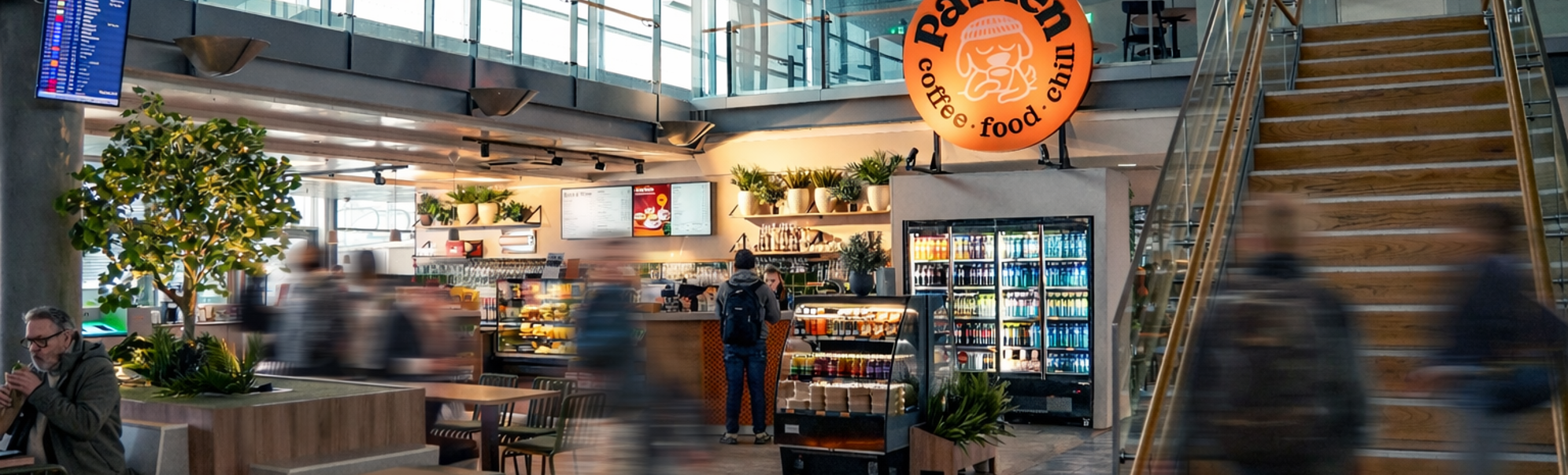 Interior of an airport café with a sign promoting spaghetti bolognese for 115 currency units; the café is named Parken and features a relaxed atmosphere with seating and plants