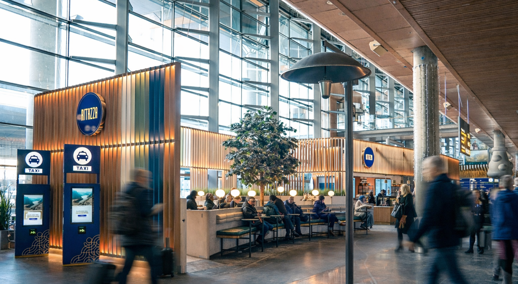Modern coffee shop interior with decorative wooden panels, hanging lights, and a large indoor tree providing a cozy atmosphere. Patrons seated at tables with coffee and snacks on display